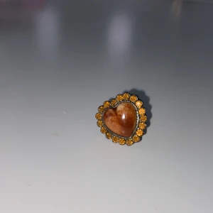 Hjärta ring med bruna stenar - Säljer en unik ring med ett hjärta i mitten omgivet av små bruna stenar. Ringen har en justerbar band för perfekt passform. Perfekt för att ge en vintage touch till din stil.