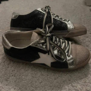 Svarta sneakers med stjärndetalj - Snygga svarta sneakers med en vit stjärna på sidan och glittriga silverdetaljer. Skorna har snörning och en unik textur på ytan som ger dem en cool look. Perfekta för att ge din outfit en extra touch.