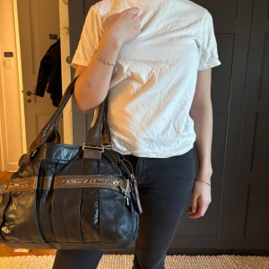 Chloé väska - Svart axelväska med bruna detaljer - Snygg svart axelväska i skinn med bruna detaljer och dragkedjor. Väskan har två handtag och flera praktiska fack. Perfekt för att bära med sig det viktigaste med stil.