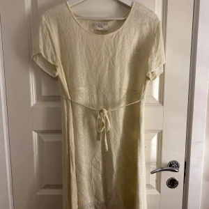 Beige klänning från Vila - Säljer en stilren beige klänning från Vila med korta ärmar och knytband i midjan. Klänningen har ett subtilt mönster och är perfekt för en avslappnad look. Passar bra till både vardag och speciella tillfällen.
