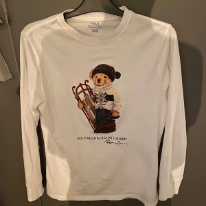 Vit långärmad tröja med björntryck från Polo Ralph Lauren - Säljer en vit långärmad tröja från Polo Ralph Lauren med ett charmigt björntryck på framsidan. Perfekt för en avslappnad och stilren look. Tröjan är i bomull och har en klassisk passform.