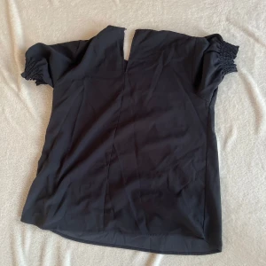 Marinblå blus med smockade ärmar - Snygg svart blus från MQ med smockade korta ärmar. Perfekt för en stilren look. Blusen har en lös passform och är tillverkad i ett mjukt material.