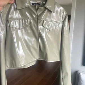 Grön glansig jacka från Gina Tricot - Snygg grön glansig jacka från Gina Tricot. Jackan är croppad med långa ärmar och har en stilren design med krage och två bröstfickor. Perfekt till en festival eller för att klä upp en basic outfit. Älskar denna men inte fått användning av den! Storlek L men passar mig som är en S då den är croppad. 