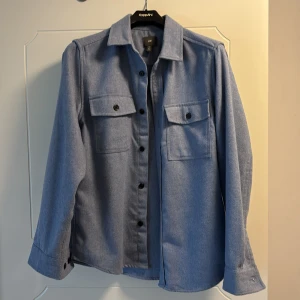 Blå overshirt från H&M - Snygg blå overshirt från H&M med långa ärmar och knappar framtill. Två praktiska bröstfickor med lock och knäppning. Perfekt för lager-på-lager-stil.