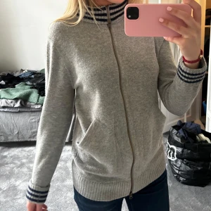 Grå kofta med dragkedja - Säljer en stilren grå Kashmir kofta med dragkedja och randiga detaljer i blått på krage och ärmslut. Kragen kan anpassas till hur du vill ha det. Perfekt för en avslappnad look. Koftan har långa ärmar och praktiska fickor framtill.