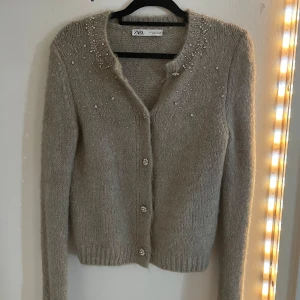 stickad kofta från Zara - Säljer en elegant grå/beige stickad kofta från Zara med pärldetaljer och glittriga knappar. Perfekt för att ge en touch av glamour till din outfit. Koftan har långa ärmar och en mjuk, bekväm passform.