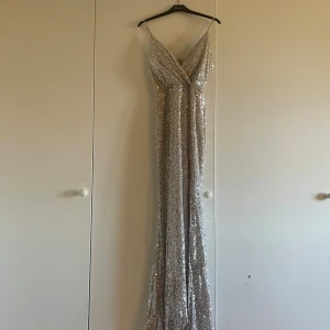 Glittrig långklänning med paljetter - Säljer en fantastisk glittrig långklänning i silver med paljetter. Klänningen har en v-ringad design och en elegant slits framtill. Perfekt för festliga tillfällen!