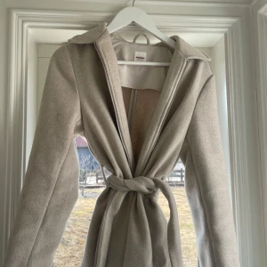 Beige kappa med bälte - Säljer en stilren beige kappa med dragkedja och knytbälte i midjan. Perfekt för höst och vår. Den har långa ärmar och en klassisk krage. Enkelt att matcha med olika outfits. Orginalpris: 699 kr