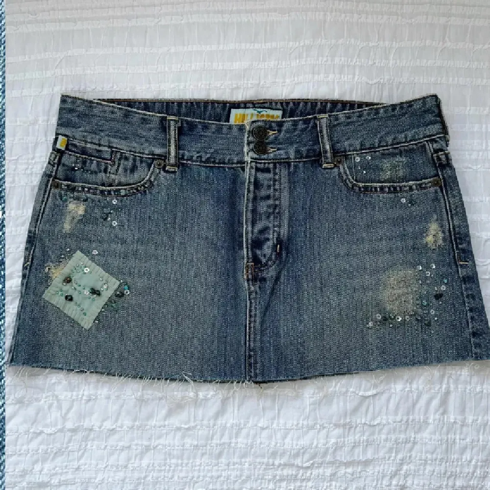Trendig jeanskjol med slitna detaljer och pärlbroderier. Kjolen har en kort längd och rå kant nedtill. Y2K, går inte att få tag på längre!!! Helt ny. Storlek 9 (27/28?). Midjemått 82 cm, framsidans längd 28 cm, baksidans längd 31 cm. Priset går inte att diskutera, använd gärna KÖP NU.. Hameet.