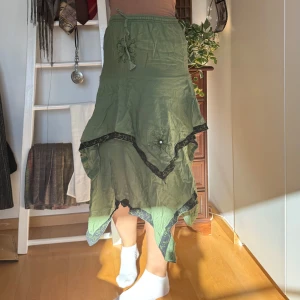 Grön kjol med asymmetrisk design - Snygg grön kjol med en unik asymmetrisk design och svarta broderade detaljer. Kjolen har en elastisk midja och dekorativa blommor. Perfekt för en bohemisk stil.