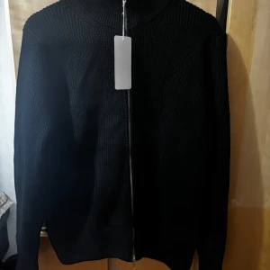Maison margiela tröja - Snygg svart stickad tröja från Maison Margiela med dragkedja framtill. Tröjan har en ribbad design och långa ärmar, perfekt för kyligare dagar. Tillverkad i Italien med hög kvalitet.