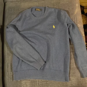 Blå tröja från Polo Ralph Lauren - Säljer en stilren blå tröja från Polo Ralph Lauren med det klassiska gula logotypbroderiet på bröstet. Tröjan har långa ärmar och ribbade muddar vid ärmslut och nederkant. Perfekt för en avslappnad och snygg look.
