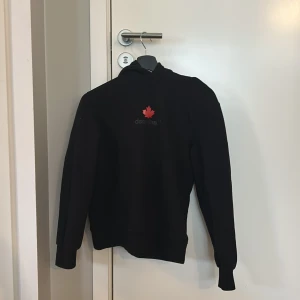Svart dsquared2 hoodie - Säljer en svart hoodie från Dsquared2 med en röd lönnlogga på bröstet. Tröjan har långa ärmar och en klassisk huva. Perfekt för en avslappnad stil.