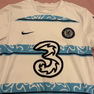 Vit och blå Chelsea fotbollströja - Säljer en vit och blå Chelsea fotbollströja från Nike med korta ärmar. Tröjan har ett unikt mönster i blått och vitt, med Chelsea FC-loggan och Premier League-märke på ärmen. Perfekt för alla Chelsea-fans! 💙