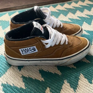 Bruna Vans Half Cab sneakers - Säljer ett par klassiska Vans Half Cab sneakers i brunt med vit snörning. Skorna har en svart insida och den ikoniska Vans-loggan på sidan. Perfekta för skate eller vardagsbruk. Skorna är knappt använda så skicket är 9/10. Dm för frågor. 