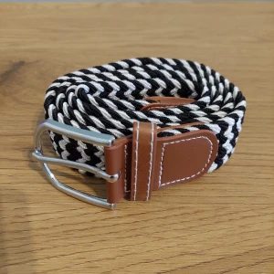 Svartvitt flätat bälte med bruna detaljer - Snyggt flätat bälte i svart och vitt med bruna läderdetaljer och silverfärgat spänne. Perfekt för att ge en stilren touch till din outfit.
