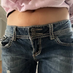 Blå jeans med slitningar - Snygga blå jeans i mycket bra skick som har slitningar och dubbla knappar i midjan. De har en lågmidjad passform och är i bootcut. Bakfickorna är dekorerade med kontrastsömmar!