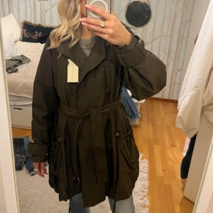 Stella McCartney x H&M Brun trenchcoat  - Snygg mörkbrun trenchcoat med bälte i midjan och knappar framtill. Jackan har långa ärmar och praktiska fickor. Perfekt för höst och vår. Jackan är också i ”typ” regnmaterial. Helt oanvänd (vintage) från Stella McCartney X H&M. 