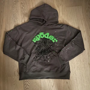 Grå hoodie med spindelnätsdesign - Cool grå hoodie från Sp5der med en unik spindelnätsdesign och gröna detaljer. Perfekt för en avslappnad stil med en touch av edge. Tillverkad i 100% bomull för extra komfort.