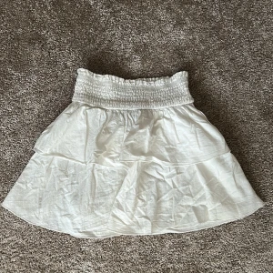 Vit kortkjol med volang - Söt vit kortkjol med volangdetaljer och elastisk midja. Perfekt för varma sommardagar. Kjolen har en luftig och bekväm passform.