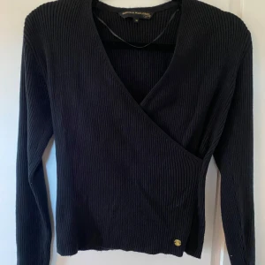 Svart omlottopp från Pernilla Wahlgren - Svart ribbad omlottopp från Pernilla Wahlgren med lång ärm och v-ringning. Toppen har en snygg guldfärgad detalj nertill och är i ett stretchigt, mjukt material som sitter skönt på kroppen. Endast använd några få gånger!