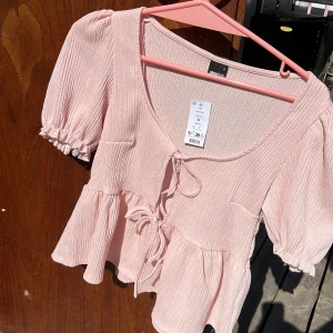 Ljusrosa blus med knyt från Gina Tricot - Supersöt ljusrosa blus från Gina Tricot med puffiga korta ärmar och volangkant. Toppen har knyt framtill och är tillverkad i ett lätt, strukturerat tyg som ger en luftig känsla. Perfekt för vår och sommar när du vill ha en romantisk vibe. Helt ny med prislapp kvar!