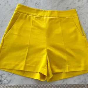Ny Gula shorts från Zara med dragkedja - Snygga gula shorts från Zara med hög midja och stilrena pressveck. Shortsen har fickor fram, dekorfickor bak och en silverfärgad dragkedja i sidan. Perfekta för dig som vill sticka ut med färg i sommar. Materialet känns mjukt och har en lätt stretch.