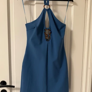 Blå kort klänning från Zara - Snygg blå kort klänning från Zara med tunna axelband och dekorativa ringdetaljer framtill. Klänningen har halterneck och en cool cut-out vid bröstet som ger en trendig look. Perfekt för dig som vill sticka ut med stilren design och unika detaljer.