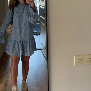 Ljusblå volangklänning med broderi från NAKD - Supersöt ljusblå kortklänning med volangkant nertill och vida, långa ärmar. Klänningen har broderade detaljer och knytning i nacken. Perfekt för dig som gillar romantisk och luftig stil. I storlek 34 från NAKD