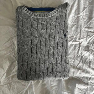 Grå kabelstickad tröja Polo Ralph Lauren - Kabelstickad grå tröja från Polo Ralph Lauren med rund halsringning och klassisk broderad logga på bröstet. Tröjan har en snygg, tidlös look och är perfekt för lager-på-lager. Mjuk och skön, passar till allt. nypris=1500 Mitt pris=700