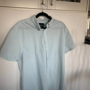 Randig kortärmad skjorta Bondelid - Fräsch kortärmad skjorta från Bondelid med tunna ljusgröna och vita ränder. Klassisk krage, knäppning framtill och broderad logga på bröstet. Perfekt för en chill och stilren look under varmare dagar.