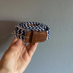 Flätat bälte - Snyggt bälte med blåvitt randigt mönster i flätad stil och detaljer i brunt skinn. Spännet är i silverfärgad metall och bältet har en modern, fräsch look som passar till jeans eller chinos. Perfekt accessoar för att lyfta din outfit.