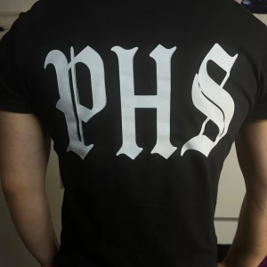 Svart PHS Support t-shirt med tryck - Svart t-shirt med stort PHS-tryck i vitt på ryggen och mindre PHS Support-logga framtill. Klassisk rund hals och korta ärmar. Materialet känns mjukt och skönt, troligtvis bomull. Snygg streetstil med tydliga kontraster i trycket.