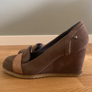Bruna kilklackade mockaloafers från Sylt - Snygga bruna loafers från Sylt med kilklack och platt sula. Skorna är i mocka med en ljusbrun läderdetalj över tån och har rund tå. Perfekta för dig som vill ha en klassisk men trendig look med lite höjd.