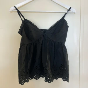 Svart broderad linnetopp med spets - Supersöt svart linnetopp med tunna axelband och broderade blommor längs nederkanten. Toppen har en luftig, lätt utställd passform och fina spetsdetaljer vid urringningen. 
