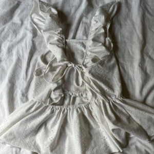 Vit broderad linnetopp med volang - Supersöt vit linnetopp med broderade detaljer och volangkant. Toppen har breda axelband med volanger och knytning i ryggen för en extra söt touch. 