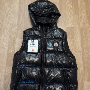 Svart dunväst från Moncler med huva - Säljer en svart, glansig dunväst från Moncler med avtagbar huva och dragkedja framtill. Västen har två fickor med dragkedja och ikonisk Moncler-logga på bröstet. Perfekt för lager-på-lager och riktigt snygg till streetwear.