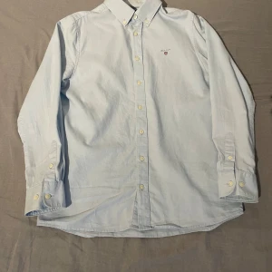 Ljusblå skjorta från Gant, 146/152 - Klassisk ljusblå skjorta från Gant med button down-krage och broderad logga på bröstet. Skjortan har långa ärmar, vita knappar och är tillverkad i mjuk bomull. Perfekt för en clean och stilren look.