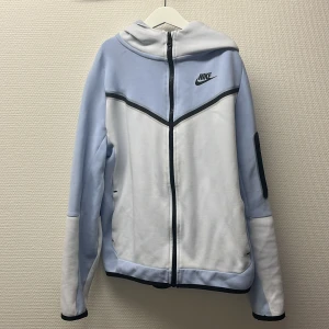 Nike tech fleece - Säljer en snygg Nike tech fleece med hoodie+byxor. Hoodien i ljusblått och vitt med svarta detaljer och dragkedja. Hoodien har huva, lång ärm och en modern design med Nike-logga på bröstet. Både byxorna och hoodien är i storlek 158-170. Perfekt för dig som gillar sportig stil.