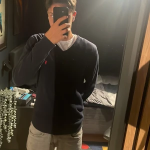 Marinblå tröja från Polo Ralph Lauren - Snygg marinblå tröja från Polo Ralph Lauren med klassisk v-ringning och röd broderad logga på bröstet. Tröjan har långa ärmar och ribbade muddar vid ärmslut och nederkant. Perfekt för en stilren och avslappnad look.