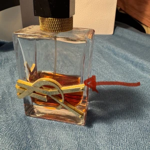 Yves Saint Laurent Libre Eau de Parfum - Snygg parfymflaska från Yves Saint Laurent med guldig logga och svart kork. Flaskan är rektangulär i genomskinligt glas och innehåller en bärnstensfärgad doft. Lyxig och modern design som sticker ut på hyllan.