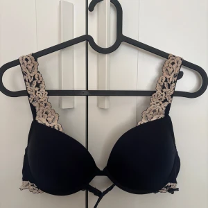 Intimissimi BH - Snygg svart bh-topp med breda axelband i beige spets och justerbara band baktill. Kuporna är vadderade och har en mjuk form. Fina spetsdetaljer längs kanterna och flera hakar för att justera omkretsen. Perfekt för dig som gillar en feminin och bekväm look.
