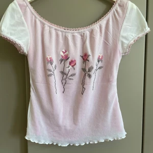 Söt rosa blus med rosor från Camera Classic - Supersöt ljusrosa blus från Camera Classic med korta vita puffärmar och broderade rosor framtill. Blusen har en söt spetskant vid halsringningen och vågig kant nertill. Perfekt för dig som gillar romantisk och gullig stil. Storlek M men liten som S