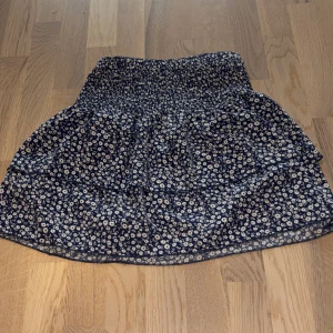 Blommig kortkjol i marinblått - Supersöt kortkjol med små vita blommor på marinblå botten. Kjolen har resår i midjan och volangig nederdel som ger en luftig och skön känsla. Perfekt för varma dagar och enkel att matcha med en enkel topp.