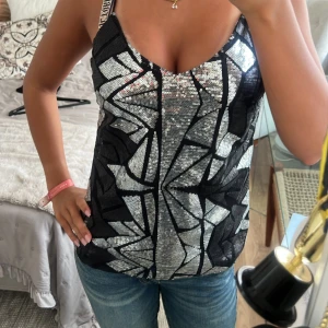 Glittrig festtopp från Noisy May - Säljer en glittrig svart och silver linnetopp från Noisy May med geometriskt mönster av paljetter. Toppen har smala axelband och v-ringning, perfekt för dig som vill sticka ut. Materialet är syntet och toppen är croppad i modellen.