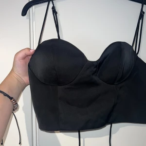 Svart korsett-topp med snörning - Säljer en svart korsett-topp med justerbara tunna axelband och snygg snörning i ryggen. Toppen har formade kupor och croppad passform, perfekt för att skapa en edgy look. Materialet är slätt och ger en stilren känsla. Passar s-l. Kuporna kan motsvara C och omkretsen är justerbar ❤️