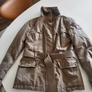 Beige parkajacka från Henri Lloyd - Snygg beige parkajacka från Henri Lloyd med bälte i midjan, höga fickor med lock och Union Jack-detalj på ärmen. Jackan har quiltat foder och hög krage, perfekt för dig som gillar klassisk stil med en twist.