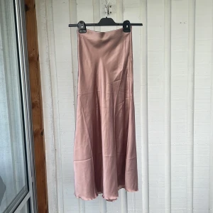 Ljusrosa satinkjol i lång modell - Superfin ljusrosa långkjol i satin med mjukt fall och lätt glansig yta. Kjolen har en enkel och stilren design utan synliga detaljer, perfekt för att skapa en chic look.