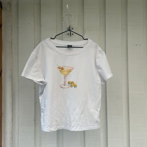 Vit t-shirt med martiniglas-tryck - Vit kortärmad t-shirt med ett färgglatt tryck av ett martiniglas och oliver på bröstet. Modellen är croppad och har en avslappnad passform med rund halsringning. Perfekt för dig som gillar roliga prints!