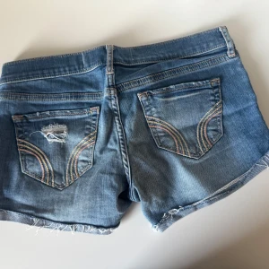 Blå jeansshorts från Hollister - Snygga blå jeansshorts från Hollister med slitna detaljer och råa kanter. Klassisk femficksmodell med broderade bakfickor och låg midja. Perfekta för sommardagar och ger en avslappnad vibe.går och rulla upp en gång till om man vill ha de lite kortare 